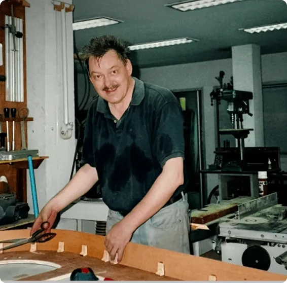A man smiling at his work bench in a workshop environment with tools and machinery visible in the background.