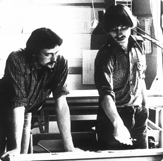 Black and white photograph of two individuals looking closely at a design schematic or document on a table.